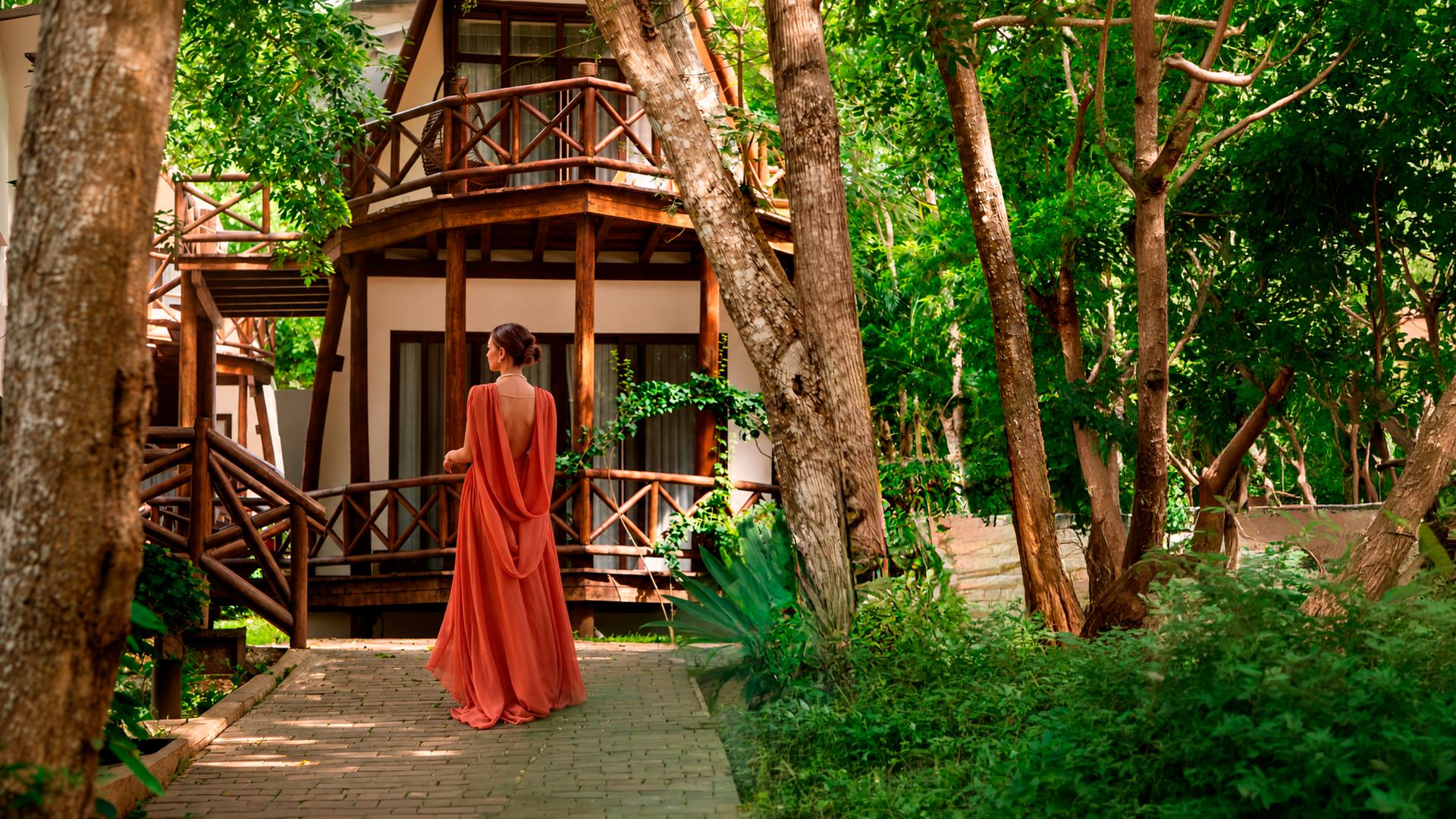 Woman in a terracotta dress walking a jungle path toward a wooden suite at MÍA Bacalar